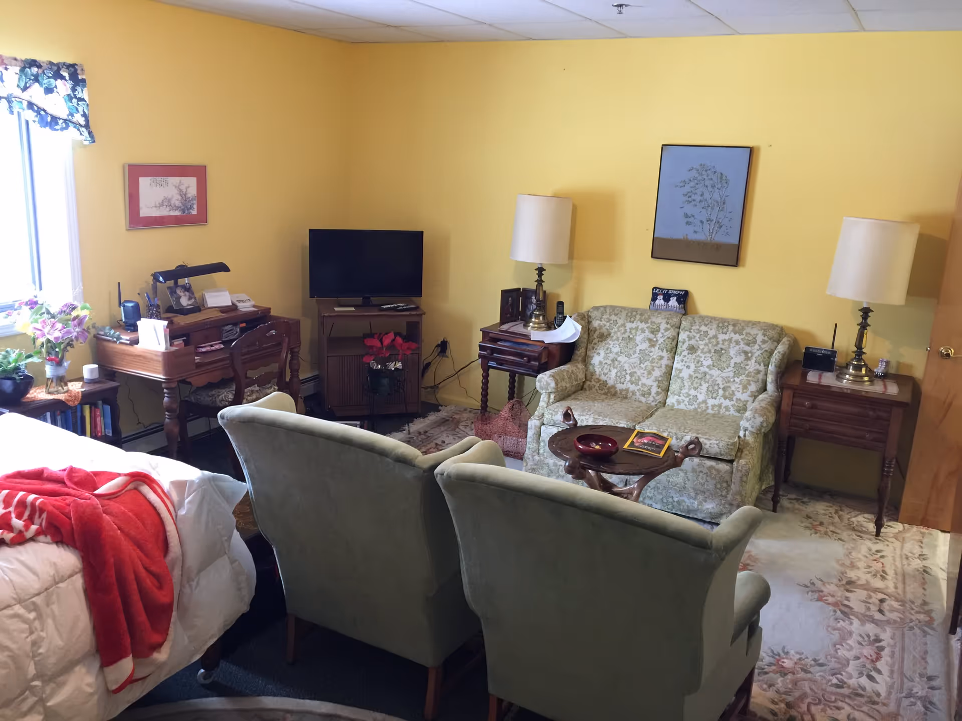 Cozy yellow-walled sitting area with a floral loveseat, two green armchairs, TV, side tables and a bed visible at left.
