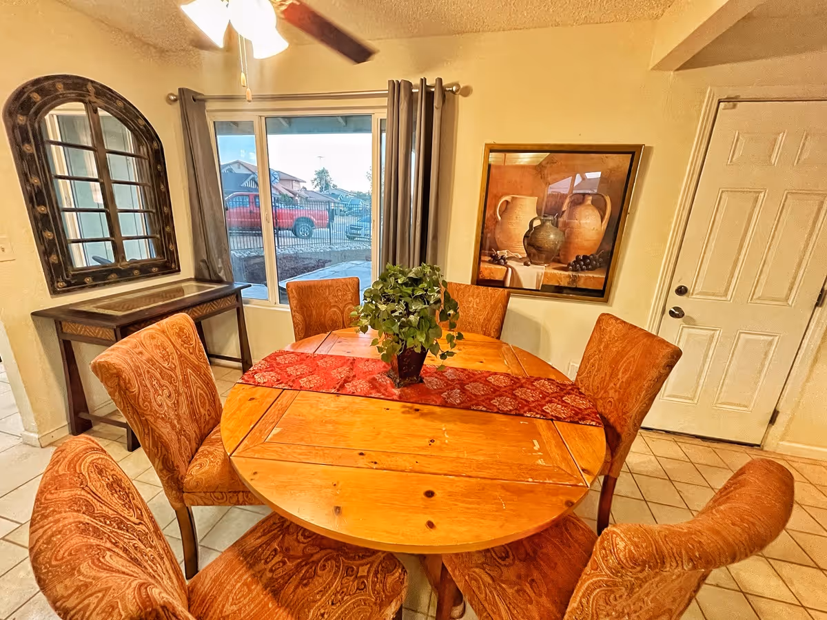 Round wooden dining table with six upholstered chairs, a red runner and a potted plant in a dining area with a window, wall art, and a door.