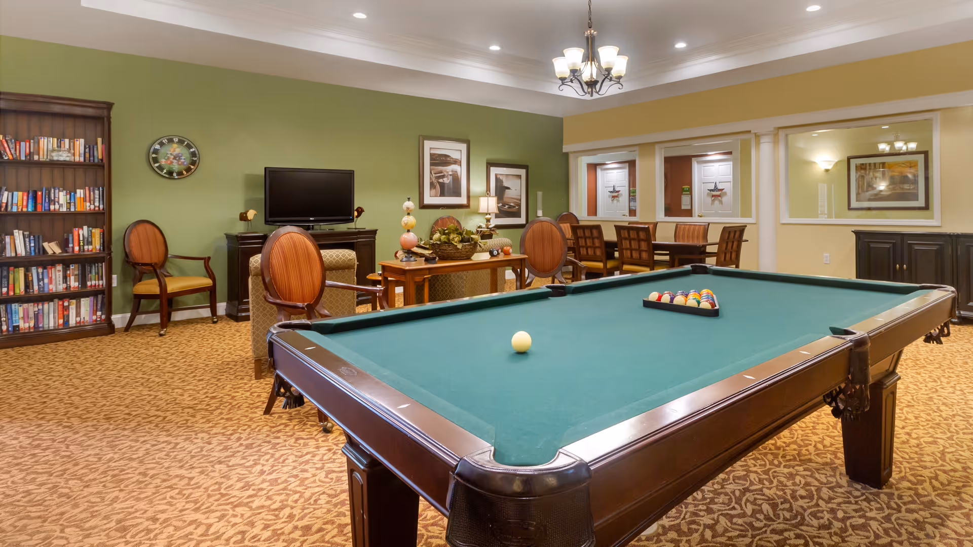 A recreational room with a pool table in the foreground, a seating area with chairs and a TV against a green wall, a bookshelf filled with books, and a dining table with chairs in the background. The room has warm lighting and carpeted flooring.