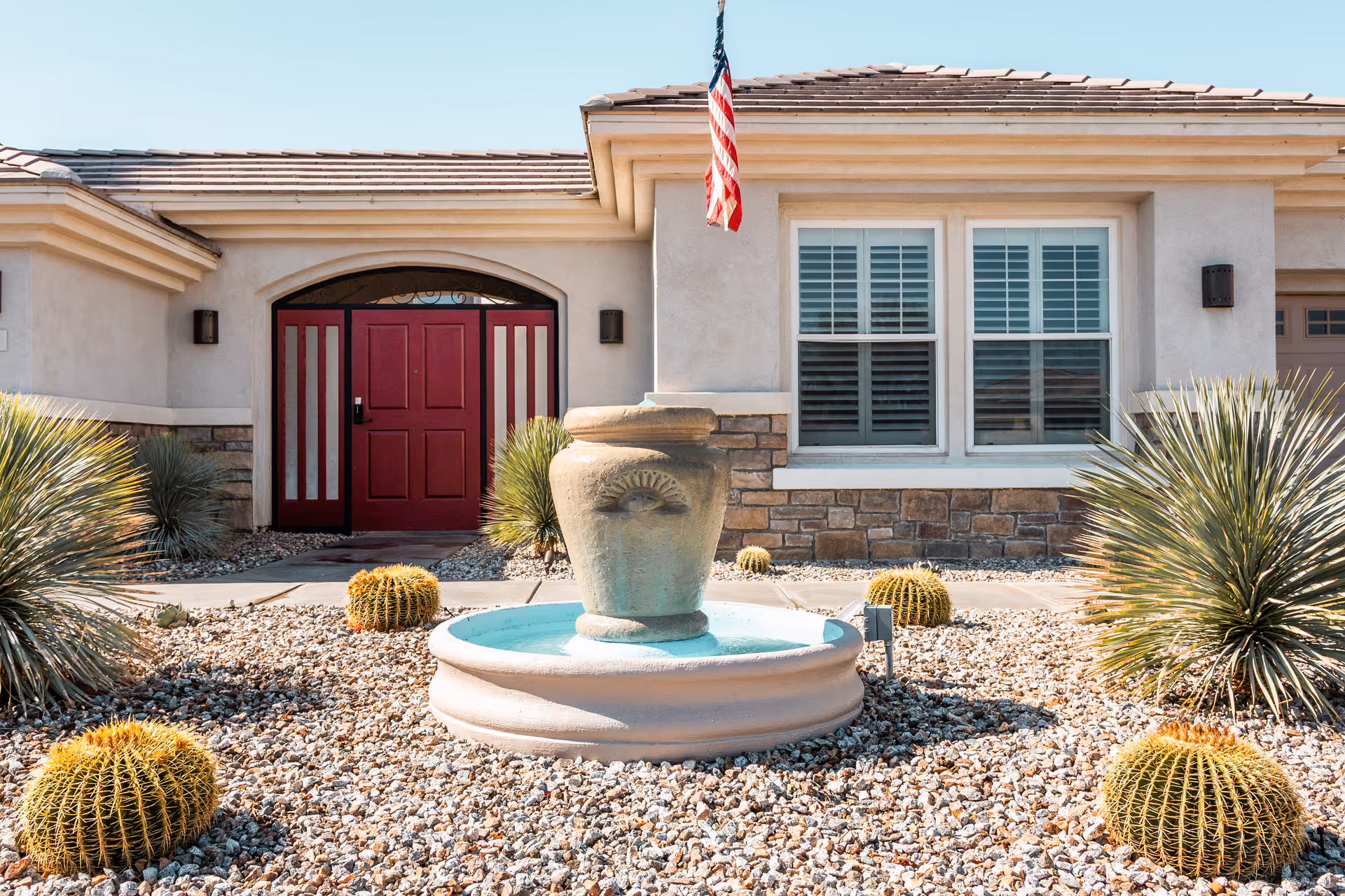 Front exterior view of a single-story building with a red double door entrance, beige walls, stone accents, and a small fountain surrounded by desert landscaping with cacti and gravel. An American flag is mounted above the fountain.