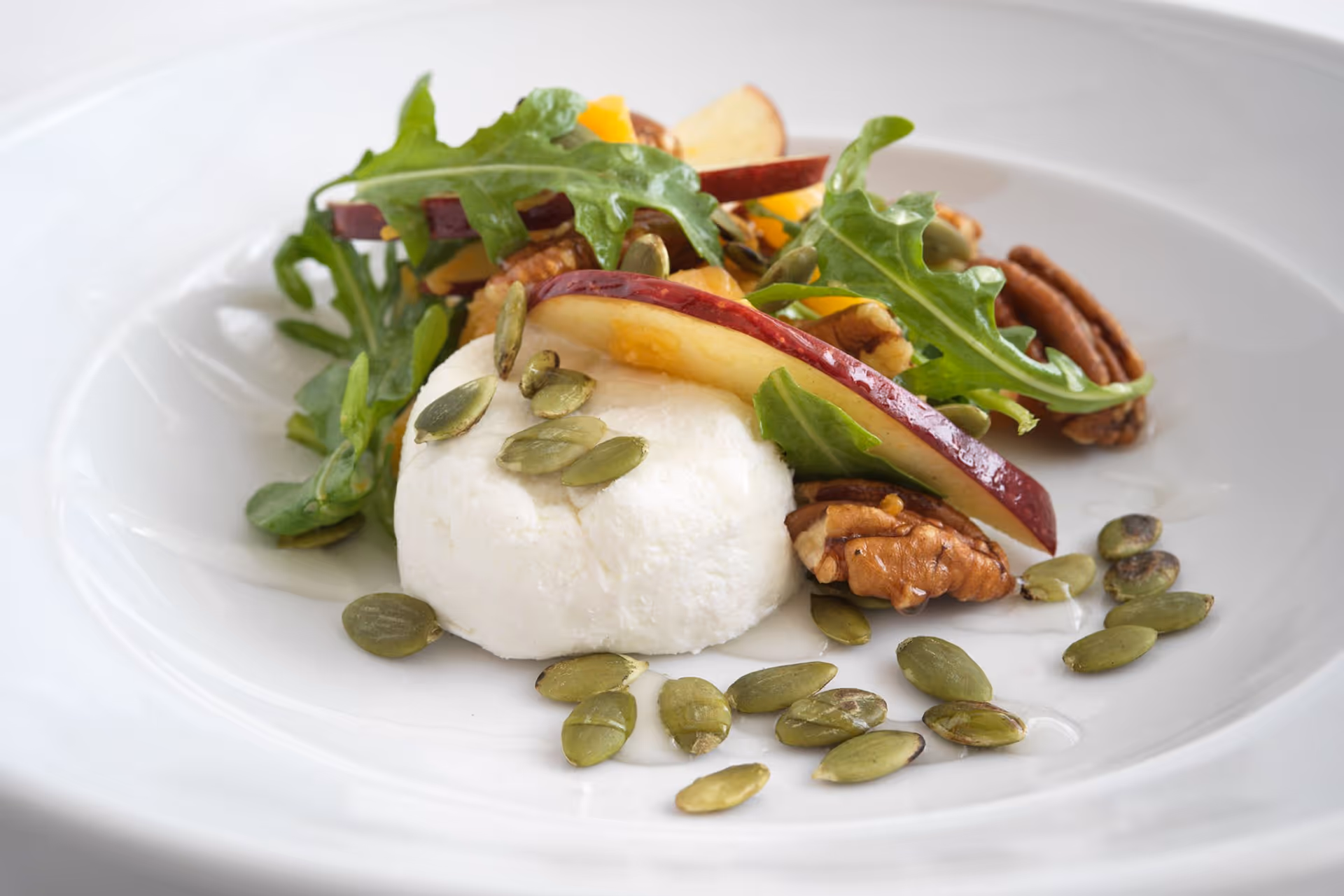 A white plate with a fresh salad consisting of arugula, sliced red apples, pecans, pumpkin seeds, and a round portion of soft white cheese.