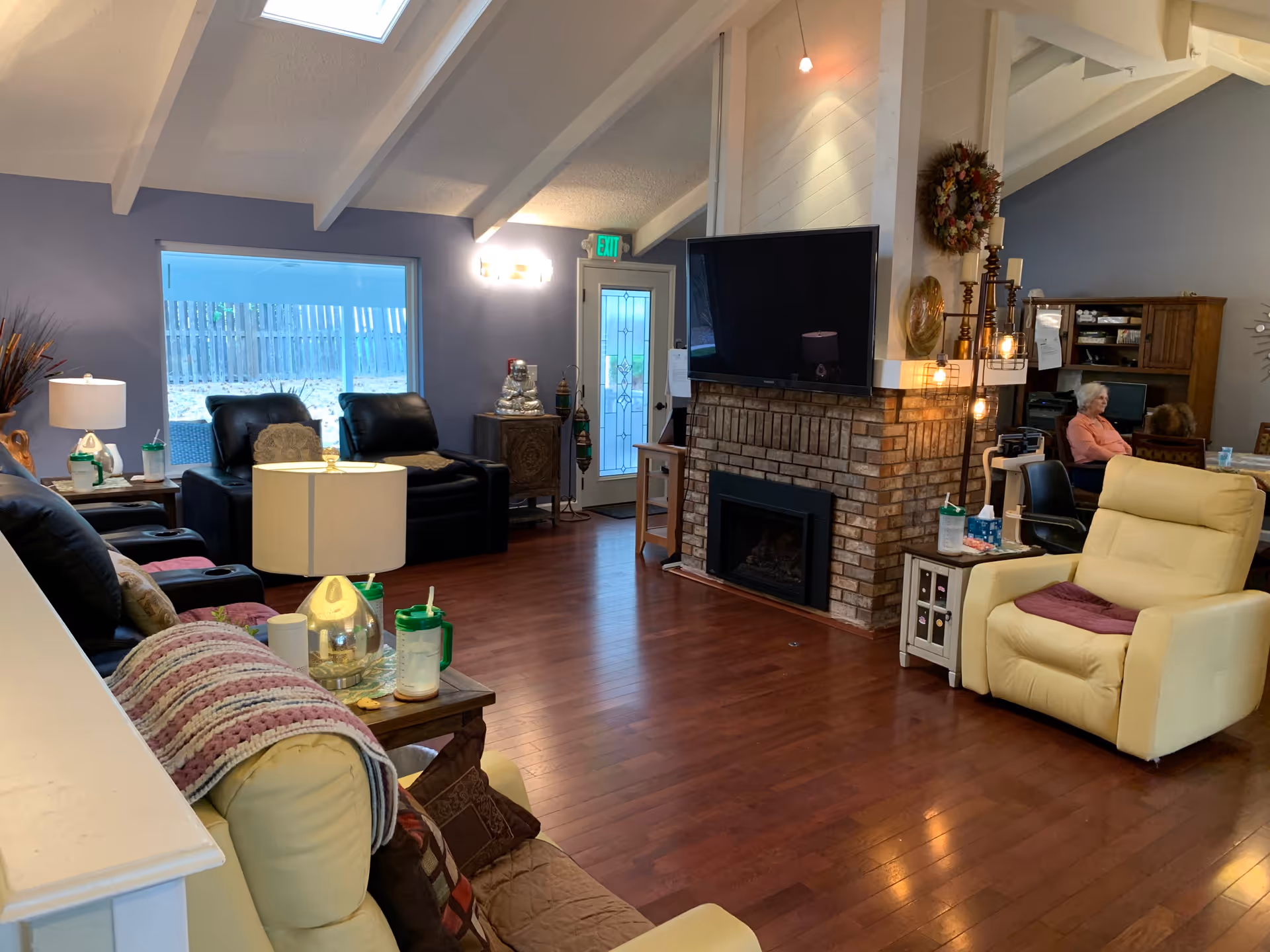 A cozy assisted living facility common area with wooden flooring, a brick fireplace with a mounted TV above it, several comfortable chairs including a yellow recliner and black leather chairs, a side table with a lamp and drinks, and a person sitting at a desk in the background. The room has a high ceiling with exposed beams and a large window letting in natural light.