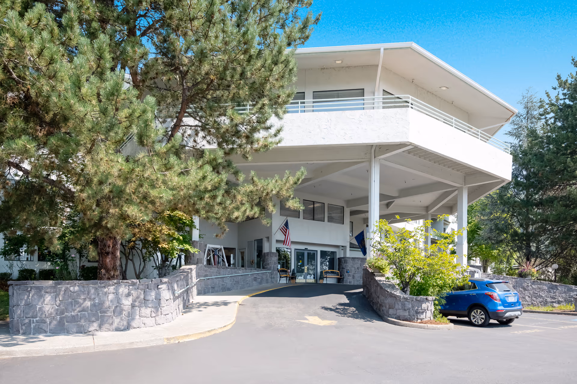 Front entrance of a multi-story white building with a covered porte-cochère, stone planters, mature trees, and a blue car parked nearby.