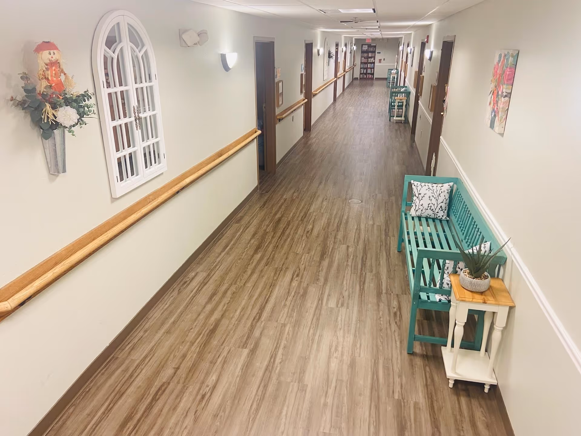 A long, well-lit hallway in a senior living facility with wood-look flooring and light-colored walls. The hallway features wooden handrails on both sides, several closed doors, and decorative elements including a turquoise bench with patterned cushions, a small side table with a plant, a wall-mounted flower arrangement, and framed artwork. At the far end, there is a bookshelf and additional seating.