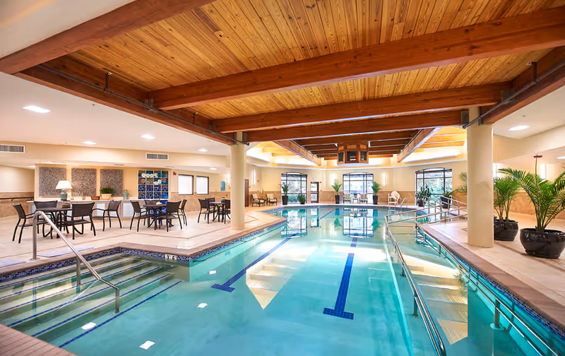 Indoor swimming pool area with clear water, steps and handrails leading into the pool, surrounded by seating areas with tables and chairs. The ceiling features wooden beams and there are large windows letting in natural light. Potted plants are placed around the pool area.