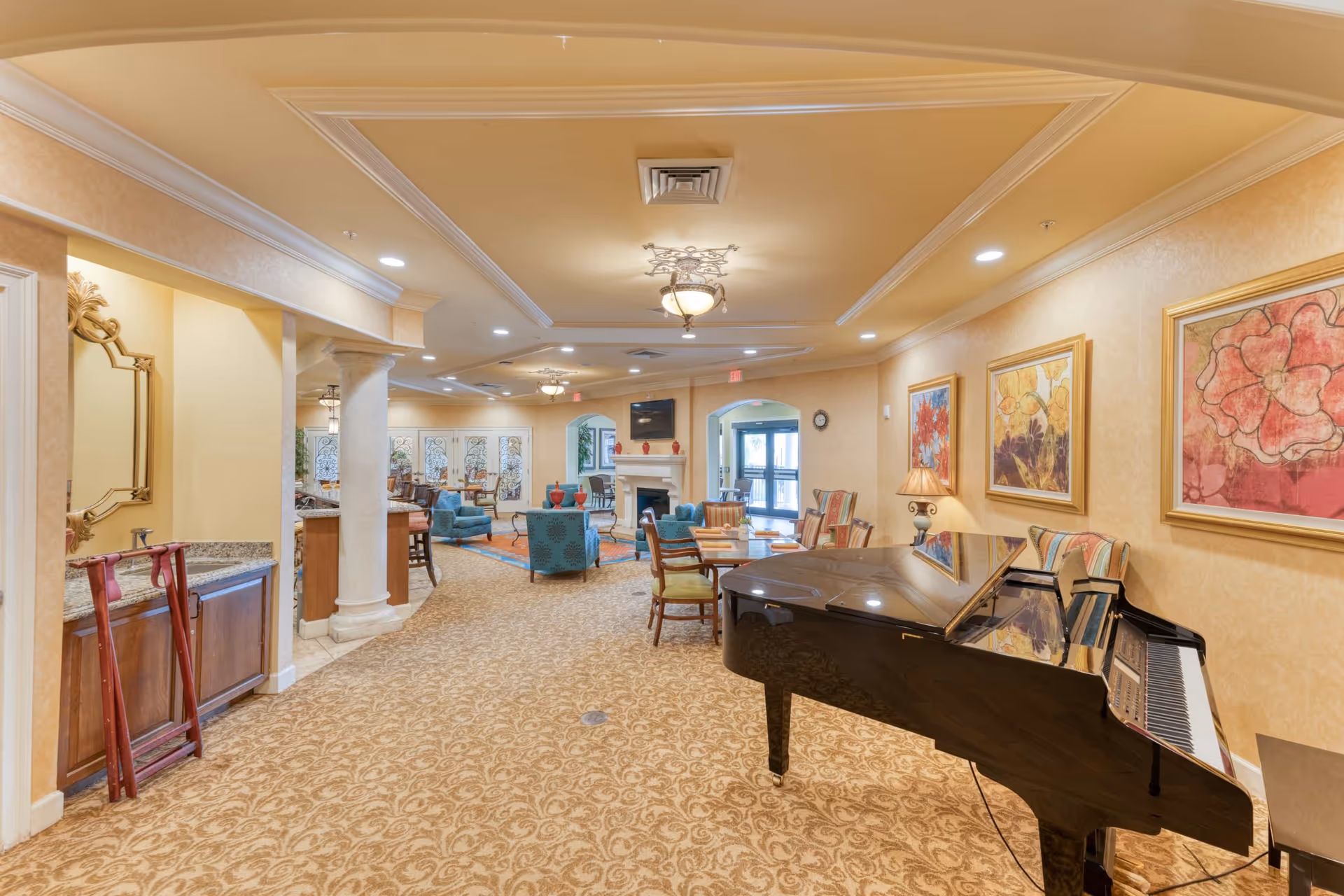 A spacious and elegantly decorated common area in a senior living facility featuring a black grand piano on the right, several seating areas with armchairs and tables, a fireplace with a TV mounted above it, and colorful floral paintings on the walls. The room has warm beige walls, patterned carpet, and ceiling lights with decorative fixtures.