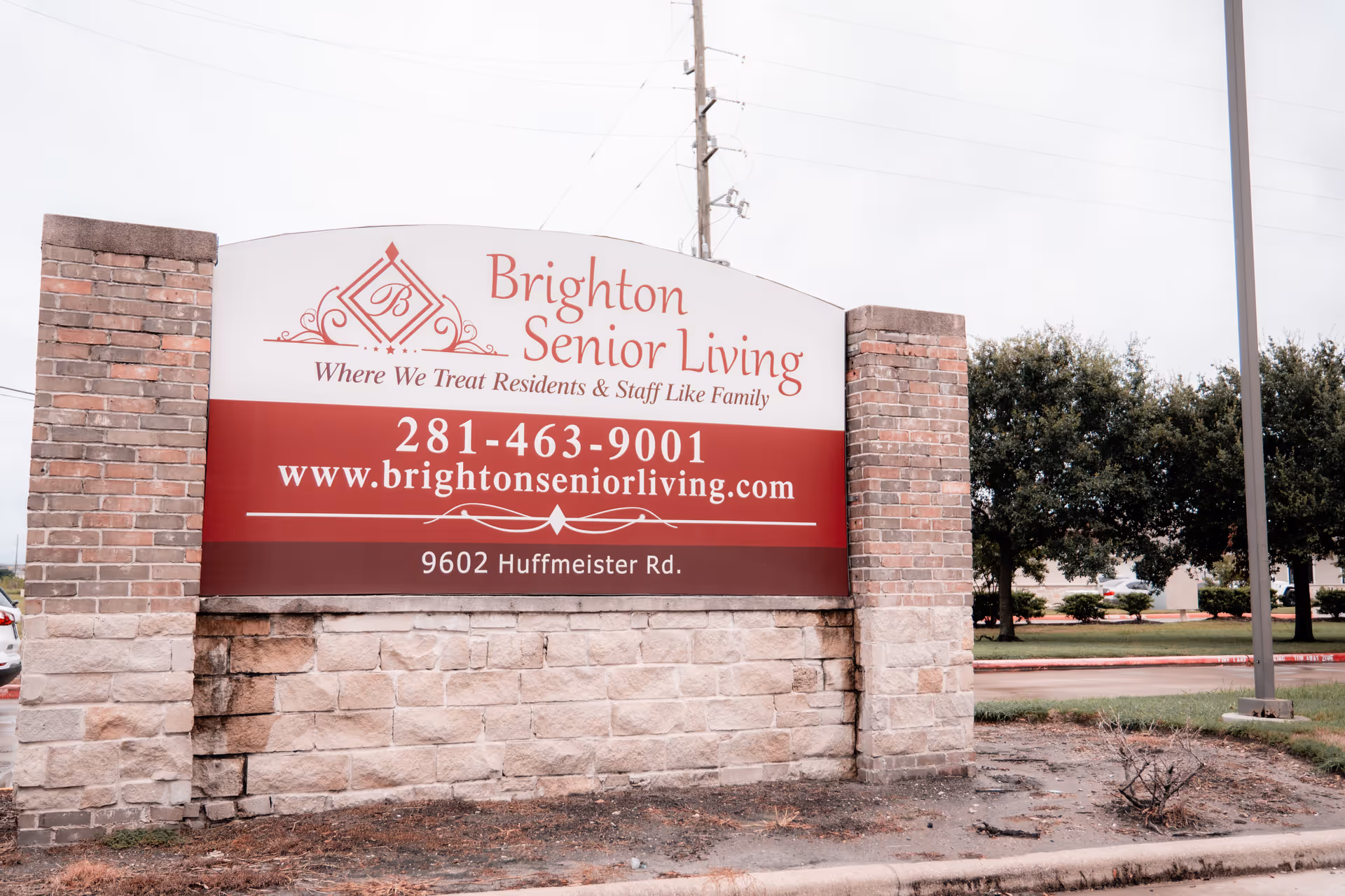Outdoor sign for Brighton Senior Living displaying the facility's name, slogan, phone number, website, and address, set between two brick pillars with trees and a parking lot in the background.