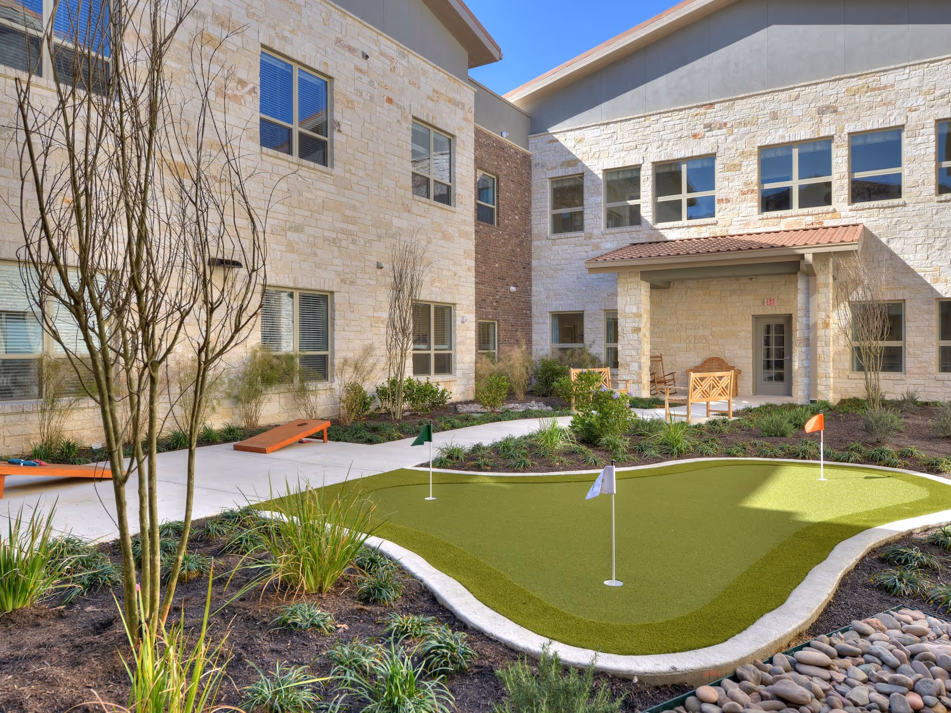 Outdoor courtyard area at Buda Oaks featuring a small putting green with three flags, surrounded by landscaped plants and shrubs. The courtyard is enclosed by a two-story building with stone and brick exterior walls, windows, and a covered seating area with wooden chairs.