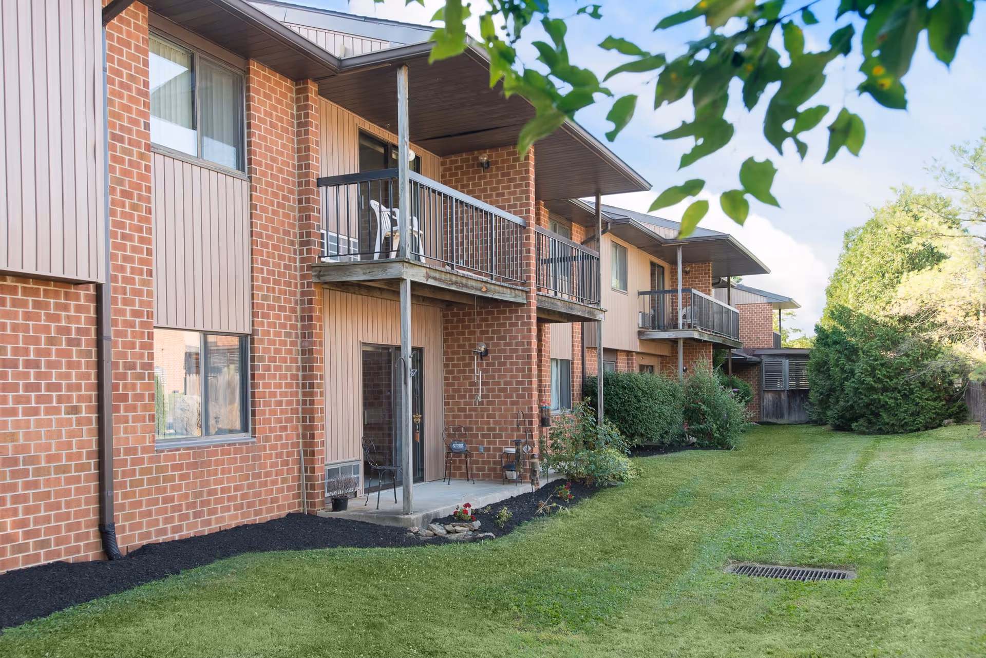 Exterior view of a two-story brick and siding building with balconies and patios overlooking a well-maintained grassy area with bushes and trees.