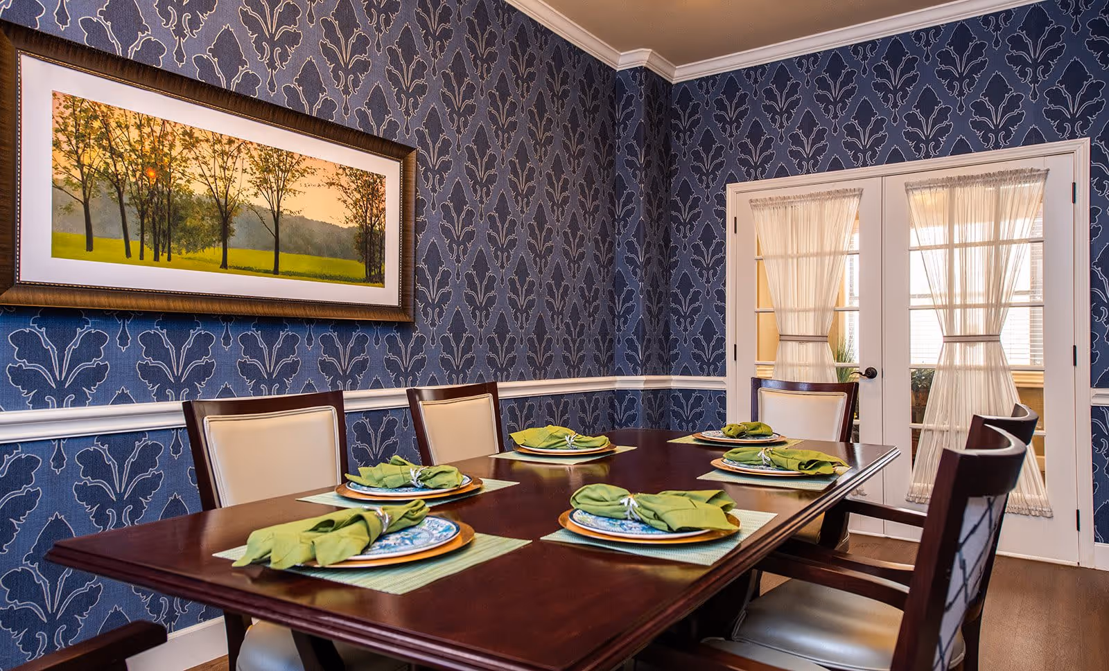 A dining room with a dark wooden table set for six people. Each place setting has a green napkin on a decorative plate with a placemat underneath. The walls are covered with blue patterned wallpaper, and there is a framed landscape painting of trees on one wall. French doors with sheer curtains are visible in the background.