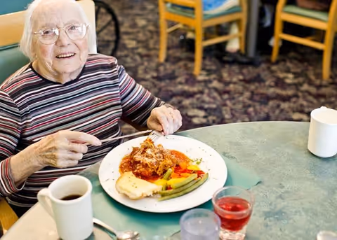 An elderly woman smiling at a dining table with a plated meal and drinks in a communal dining area.