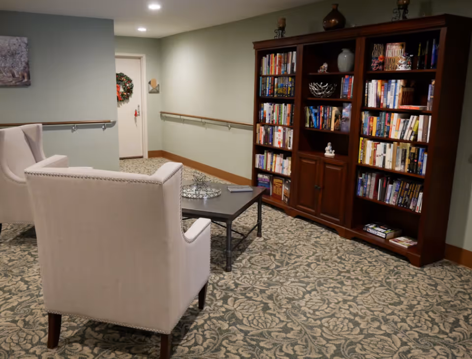 A cozy reading nook in a senior living facility featuring two beige upholstered armchairs facing a dark wooden coffee table. Behind the table is a large wooden bookshelf filled with books and decorative items. The room has patterned carpet flooring, light green walls with wooden trim, and handrails along the walls. A door with a holiday wreath is visible in the background.