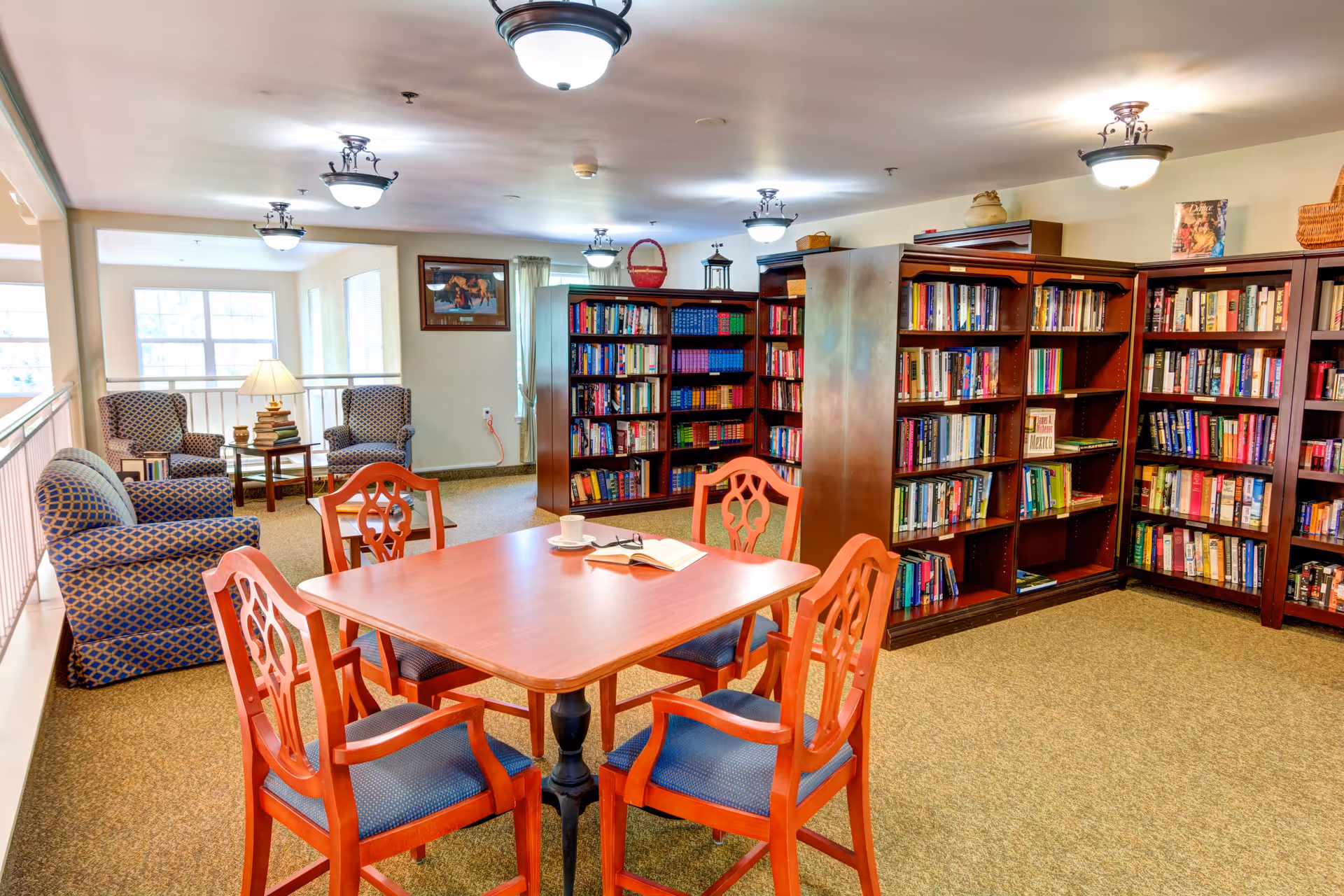 A cozy library room with wooden bookshelves filled with books along the walls. In the center, there is a wooden table with four chairs, an open book, and a cup on it. In the background, there are two armchairs with a small table and lamp between them near a window, providing a comfortable reading area. The room is well-lit with ceiling lights and has carpeted flooring.