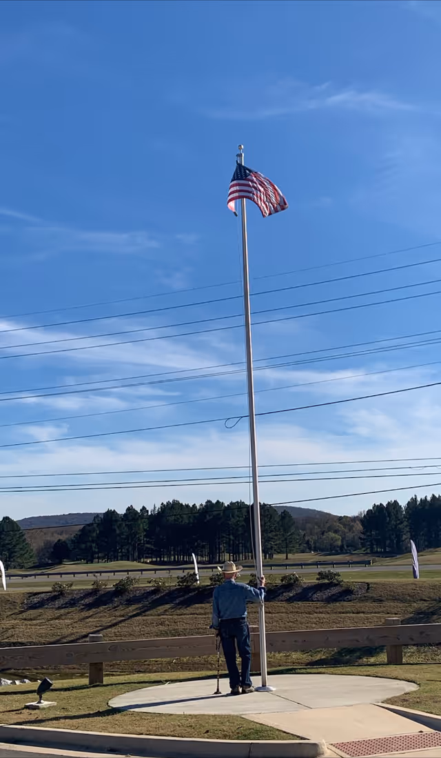 An elderly man wearing a hat and using a cane stands next to a tall flagpole with an American flag waving at the top. The scene is outdoors with a clear blue sky, trees, and hills in the background.
