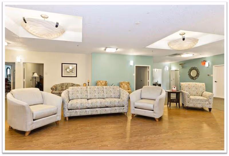 Spacious senior living common room with a sofa, several armchairs, side tables, and overhead lighting on a hardwood floor.