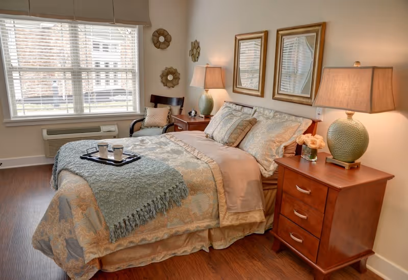 A cozy bedroom with a neatly made bed featuring patterned bedding and a light blue throw blanket. There are two wooden nightstands on either side of the bed, each with a green ceramic lamp and one with a small vase of flowers. Two framed mirrors hang above the bed. A wooden chair with a cushion is placed near a large window with blinds, and the room has wooden flooring.