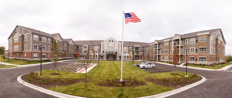 Wide exterior view of a large three-story senior living facility with beige and brick facade, multiple windows, and balconies. A well-maintained lawn with small trees and flower beds is in front, along with a flagpole flying the American flag. The driveway and parking areas surround the building under a cloudy sky.