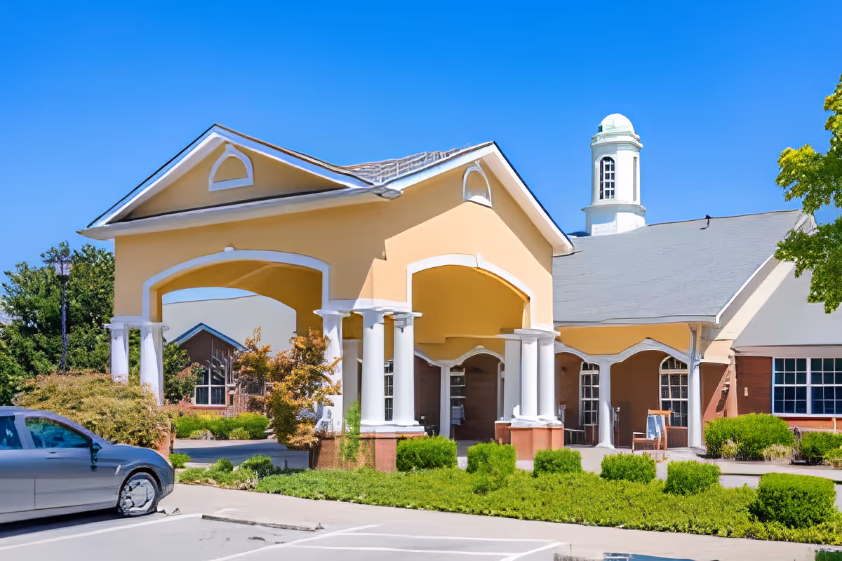 Exterior view of a senior living facility building with a covered entrance supported by white columns, surrounded by green bushes and trees under a clear blue sky. A silver car is parked near the entrance.