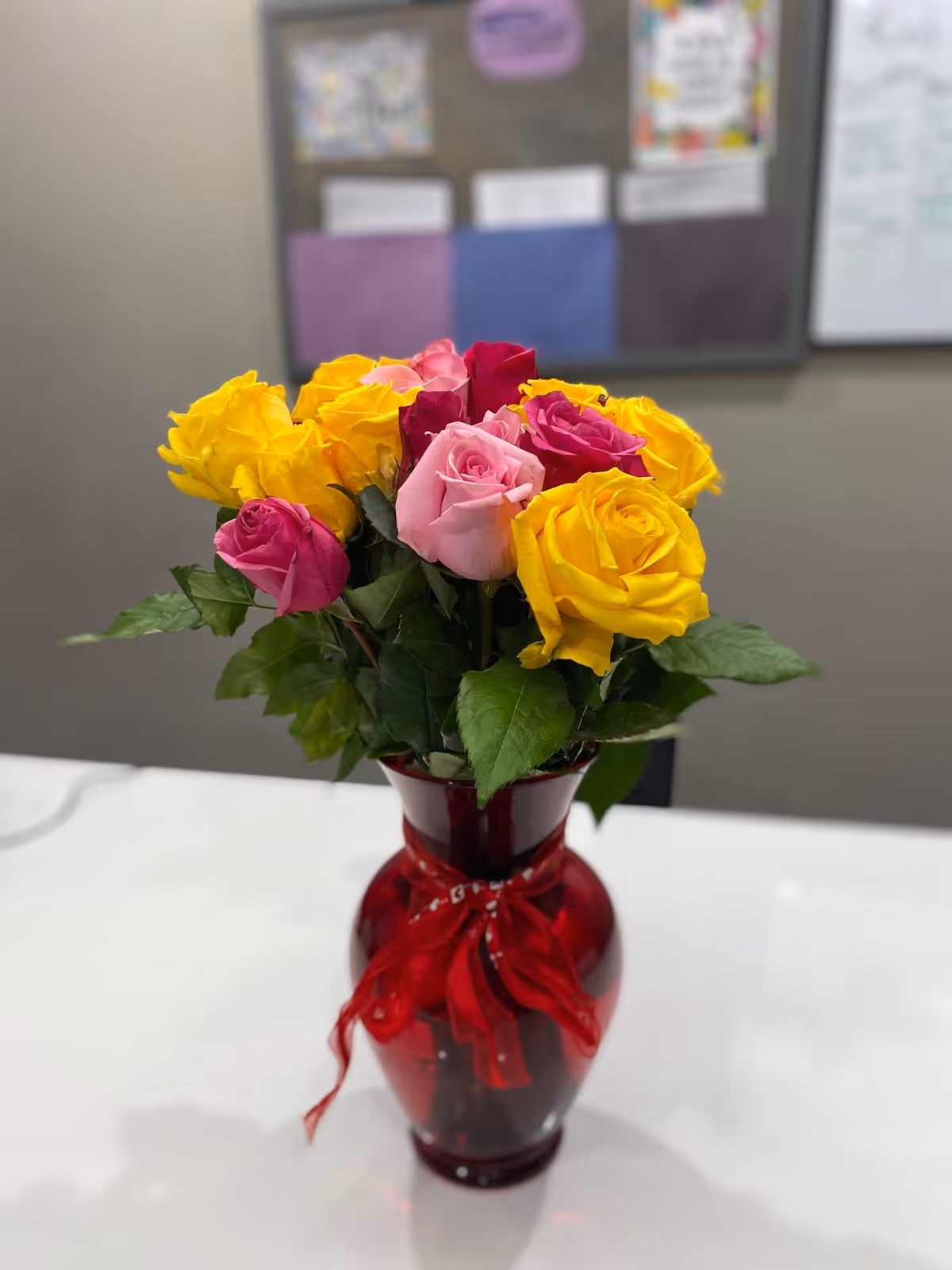 A bouquet of vibrant yellow, pink, and red roses arranged in a red glass vase with a red ribbon tied around it, placed on a white table. In the background, there is a blurred bulletin board with various papers and notes pinned on it.
