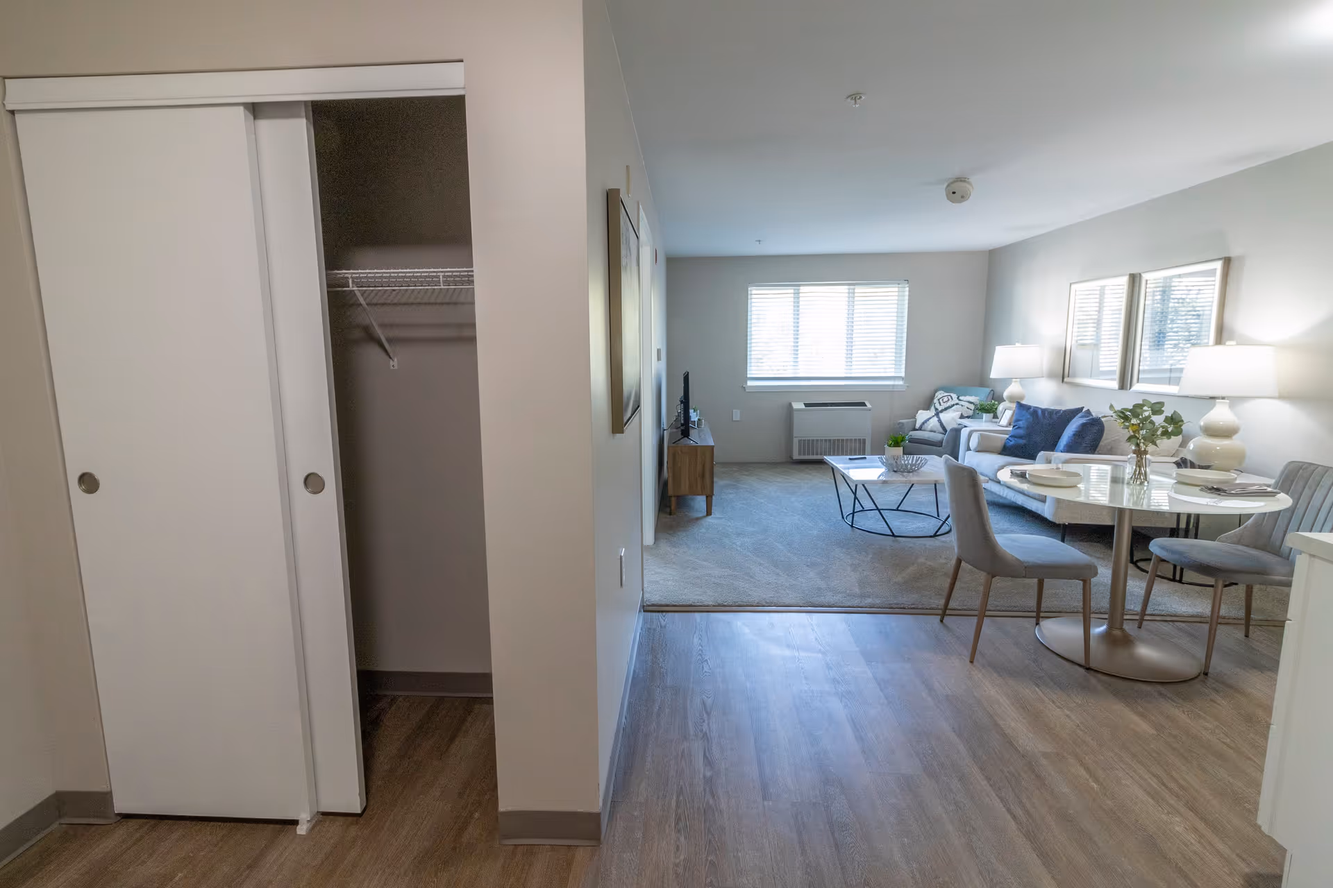 Interior view of a senior living apartment showing an open closet with sliding doors on the left, and a living room area with a sofa, coffee table, TV, and a small dining table with chairs on the right. The room has wood flooring and a window with blinds letting in natural light.