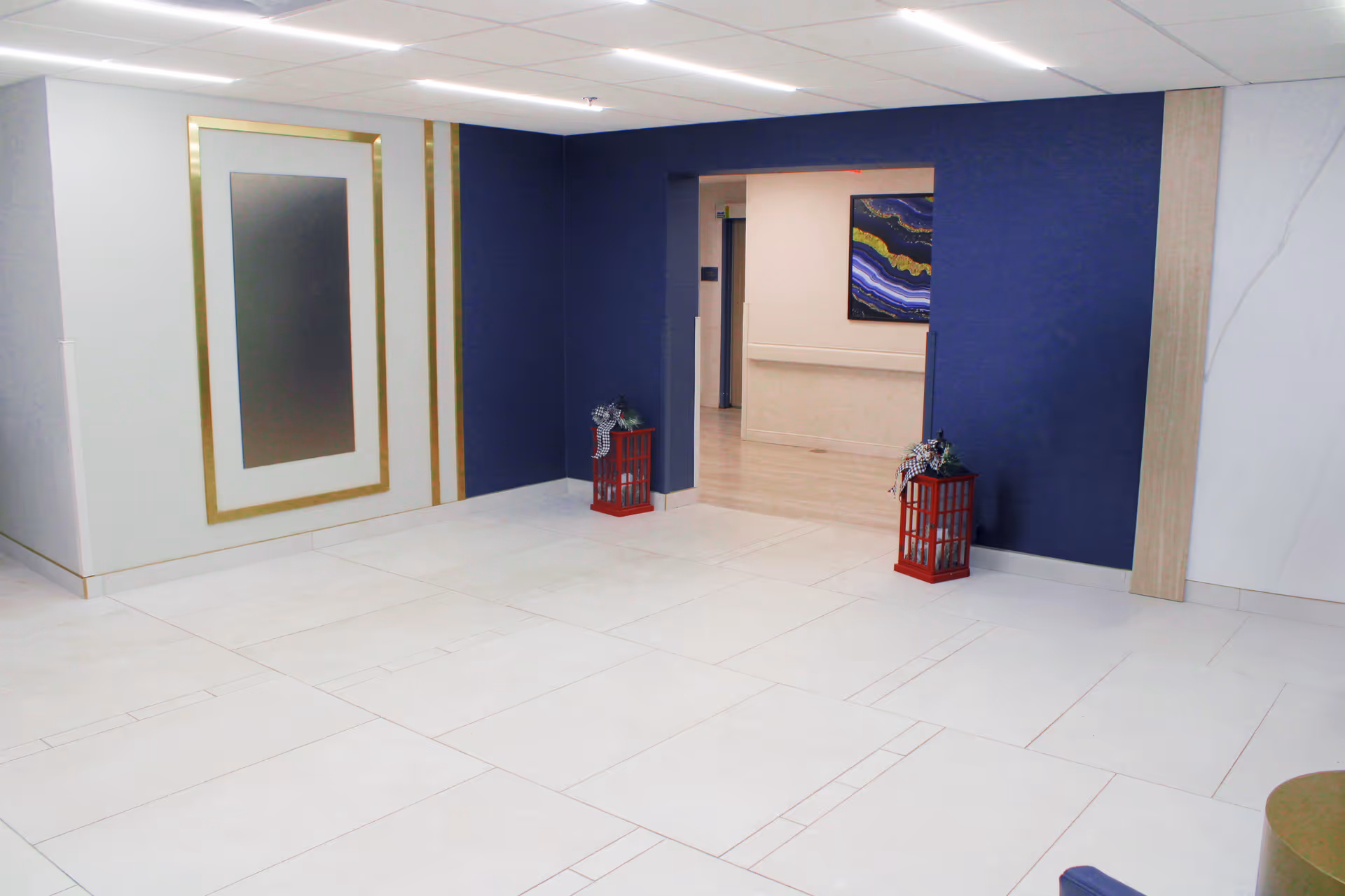 An empty interior hallway area with white tiled floors and a dark blue accent wall. Two red decorative lanterns with plants and ribbons are placed on the floor near the entrance to another hallway. The walls have white and gold trim, and a framed abstract artwork is visible in the adjacent hallway.