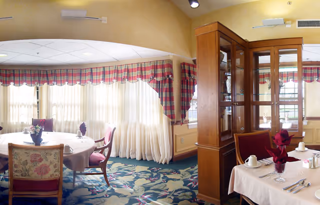 Bright, traditionally decorated dining room with tables set for meals, large windows with plaid valances, floral carpet, and wooden display cabinets.