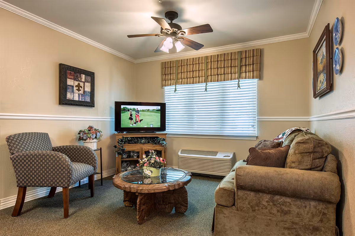 A cozy living room with a beige sofa, a patterned armchair, a round wooden coffee table with a glass top and a flower pot, a TV on a wooden stand showing a golf scene, a window with blinds and a striped valance, and wall decorations including framed pictures and decorative plates.