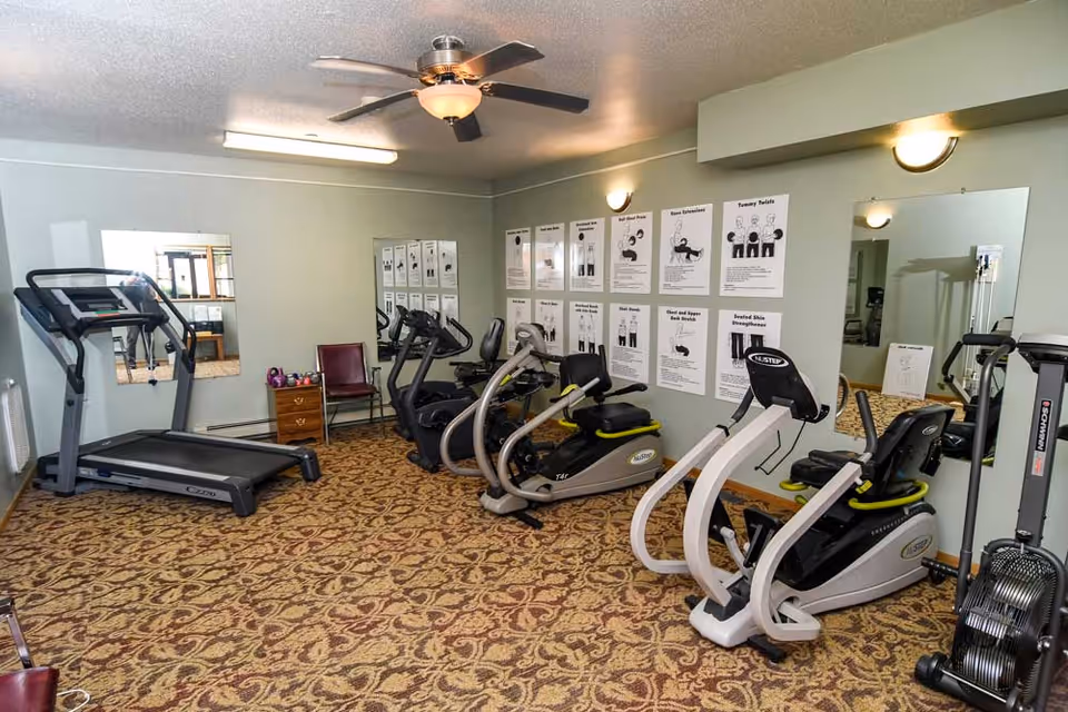 Small fitness room with treadmills, recumbent bikes, mirrors, and exercise instruction posters on the wall.