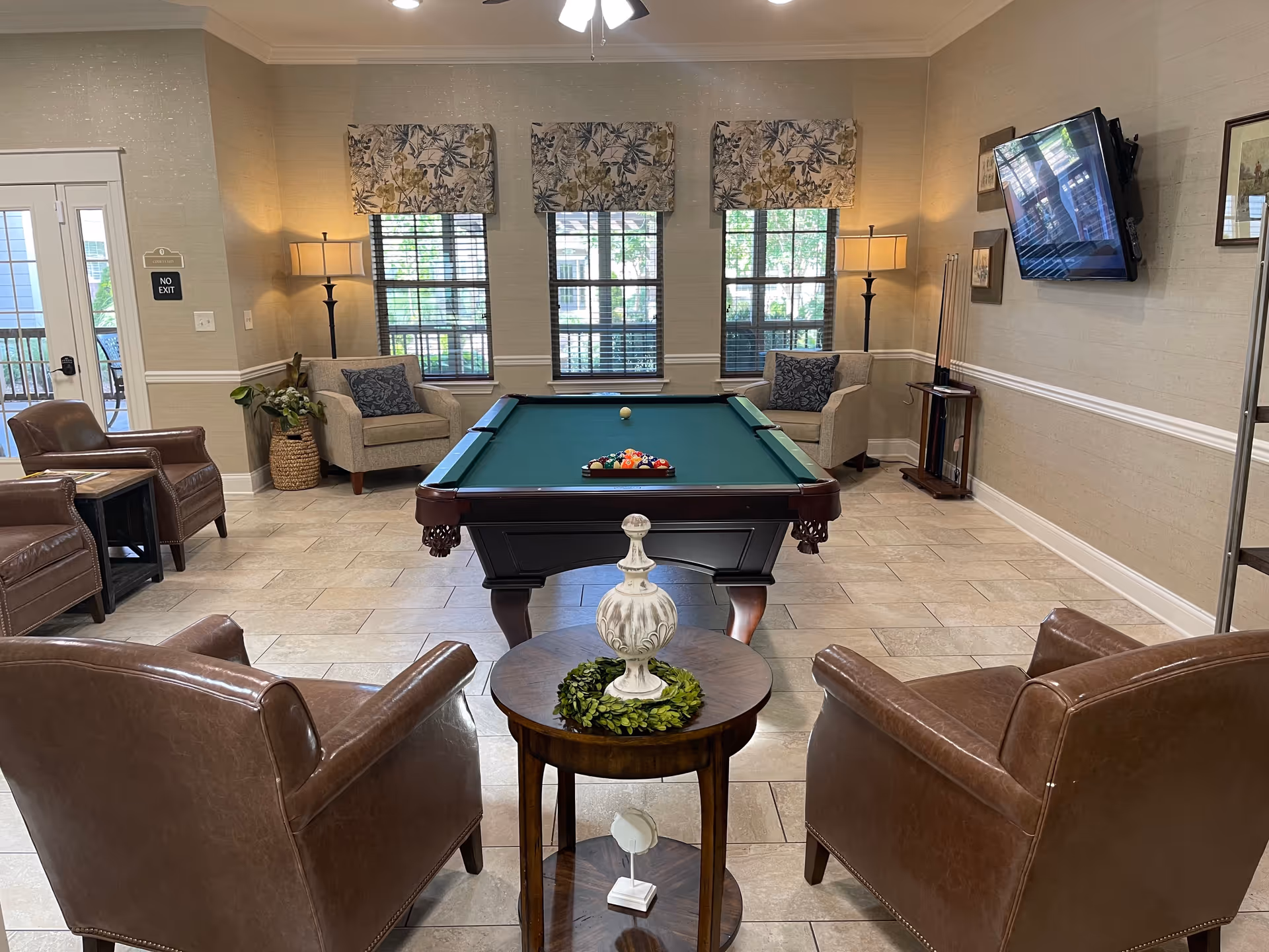 A cozy common room featuring a pool table in the center with a full rack of billiard balls and a cue ball. Surrounding the pool table are comfortable leather and fabric armchairs, two floor lamps, and a small round table with a decorative white urn and greenery. Three windows with floral valances allow natural light into the room, and a flat-screen TV is mounted on the wall to the right.