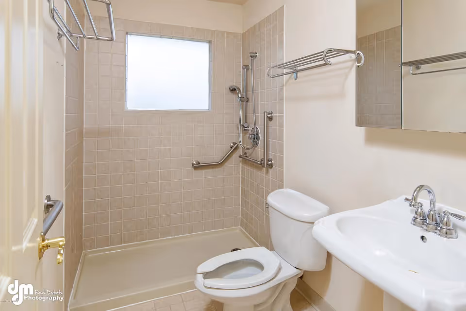 A clean bathroom featuring a walk-in shower with beige tiles and grab bars, a white toilet with an open lid, a white sink with a chrome faucet, a mirrored medicine cabinet, and a towel rack above the toilet. There is a frosted window above the shower area allowing natural light in.