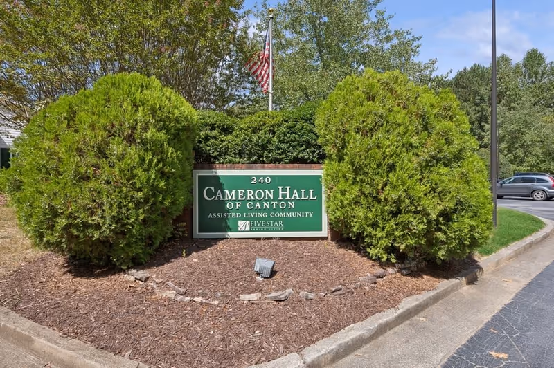 Outdoor view of a green sign for Cameron Hall of Canton, an assisted living community, surrounded by bushes and trees with an American flag in the background.