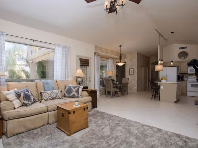 A spacious and well-lit living area featuring a beige sofa with patterned cushions, a wooden chest used as a coffee table, and a side table with a lamp. The room opens into a dining area with a glass-top table and four chairs, and a kitchen with an island, bar stools, and modern appliances. Large windows with white curtains allow natural light to fill the space.
