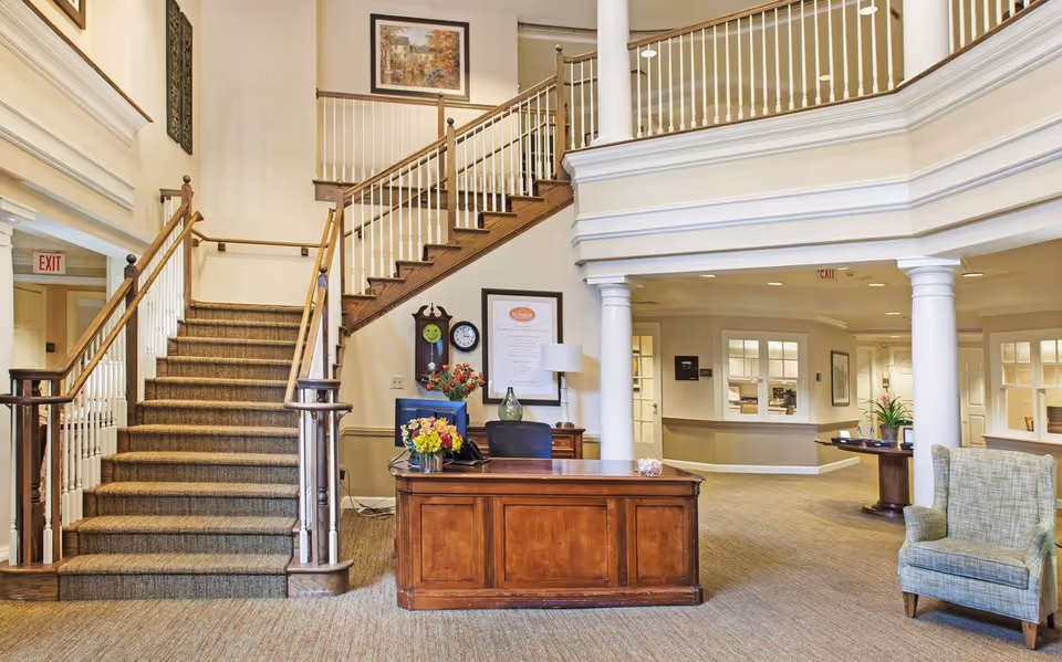 Reception lobby with a wooden desk, carpeted staircase with white spindles, columns, and seating.