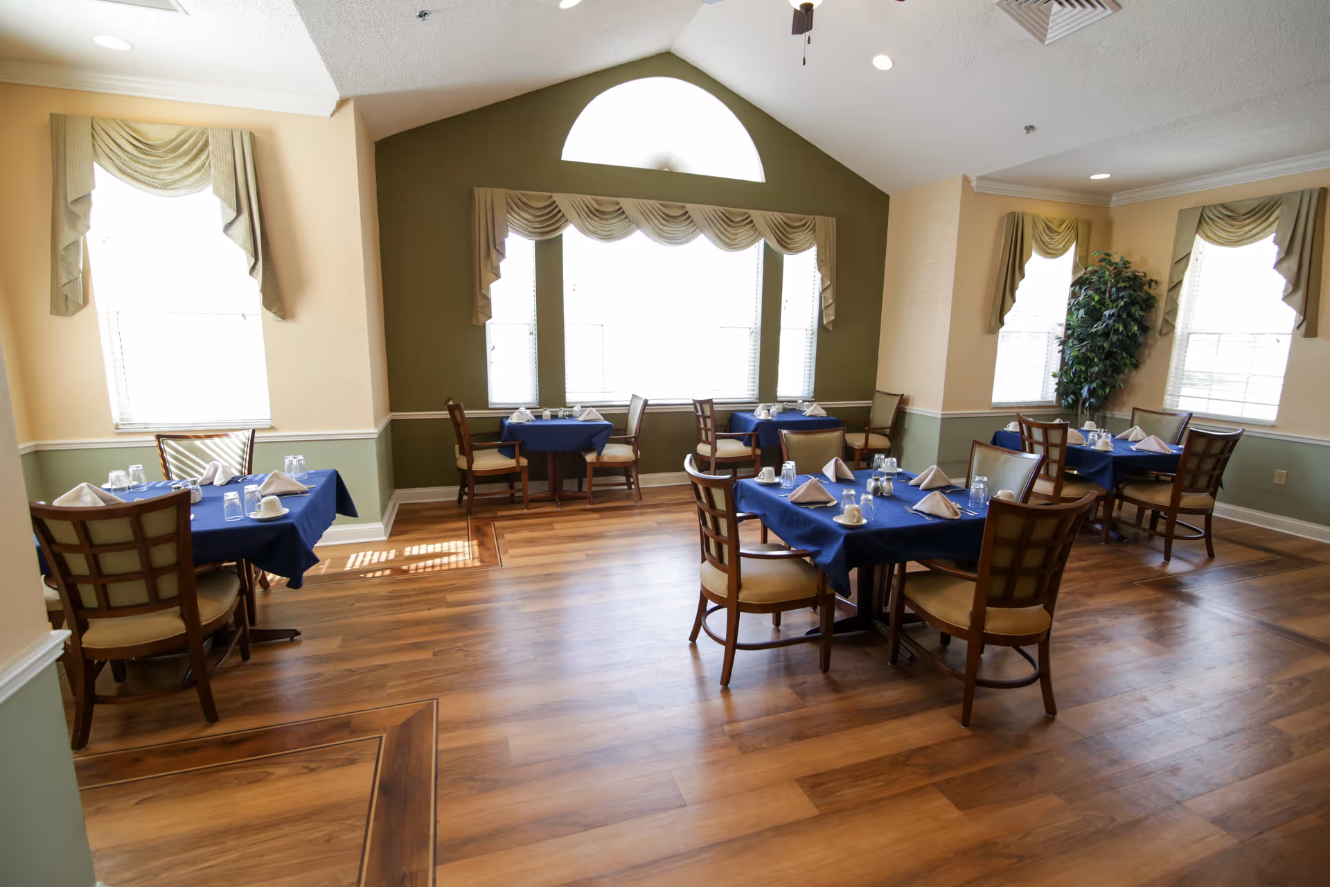 A bright dining room with wooden floors and several tables covered with blue tablecloths. Each table is set with white napkins, cups, and glasses. The room has large windows with beige curtains and a high ceiling with a ceiling fan. There is a green accent wall and a potted plant in the corner.