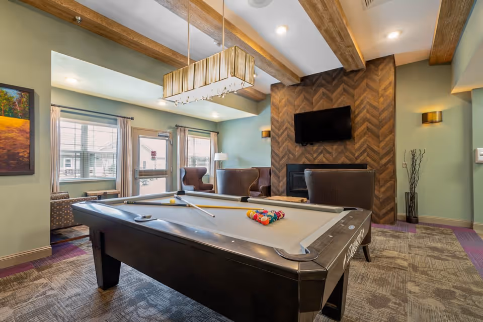 A recreational room with a pool table in the center, a cue stick resting on it, and a set of billiard balls arranged for a game. The room features a modern chandelier hanging above the pool table, exposed wooden ceiling beams, a wall-mounted flat-screen TV above a fireplace with a chevron wood pattern, and comfortable seating including armchairs near large windows with curtains.