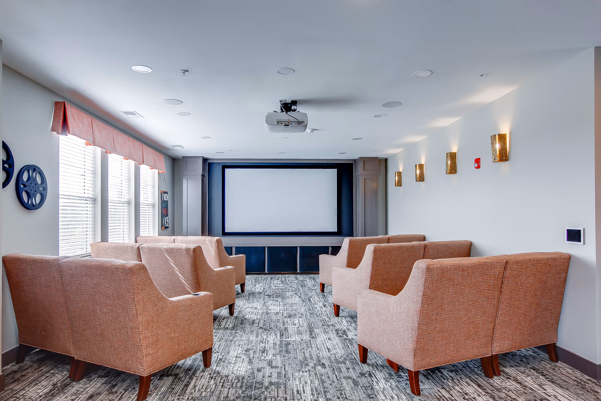 A small theater room with six beige upholstered armchairs arranged in two rows facing a large blank projector screen. The room has a patterned carpet, white walls with modern wall sconces, and three windows with blinds and a pink valance on the left side. A ceiling-mounted projector is centered above the seating area.