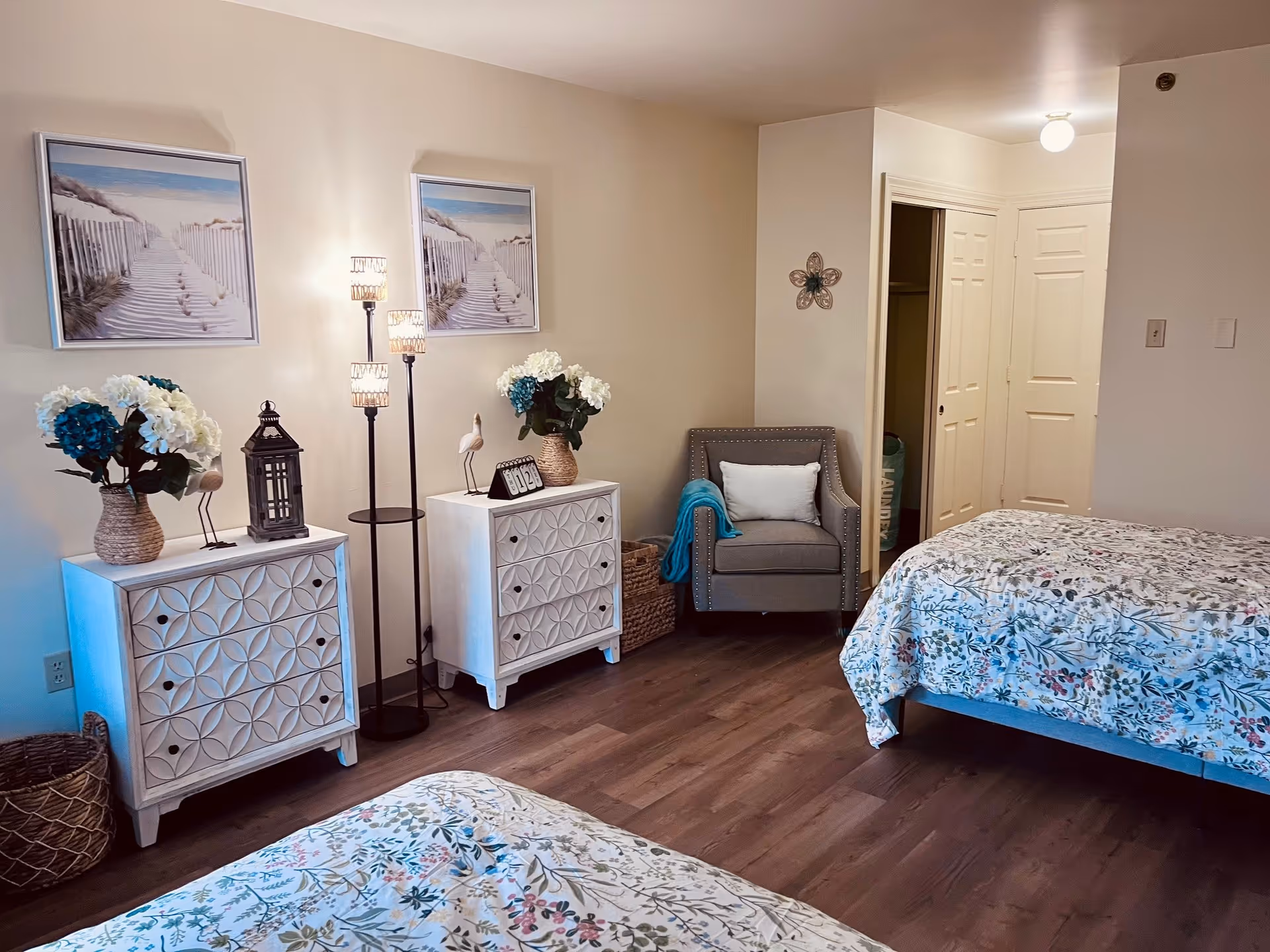 Well-lit bedroom with two beds in floral bedding, matching white decorative dressers, a gray armchair, floor lamp, and coastal wall art.