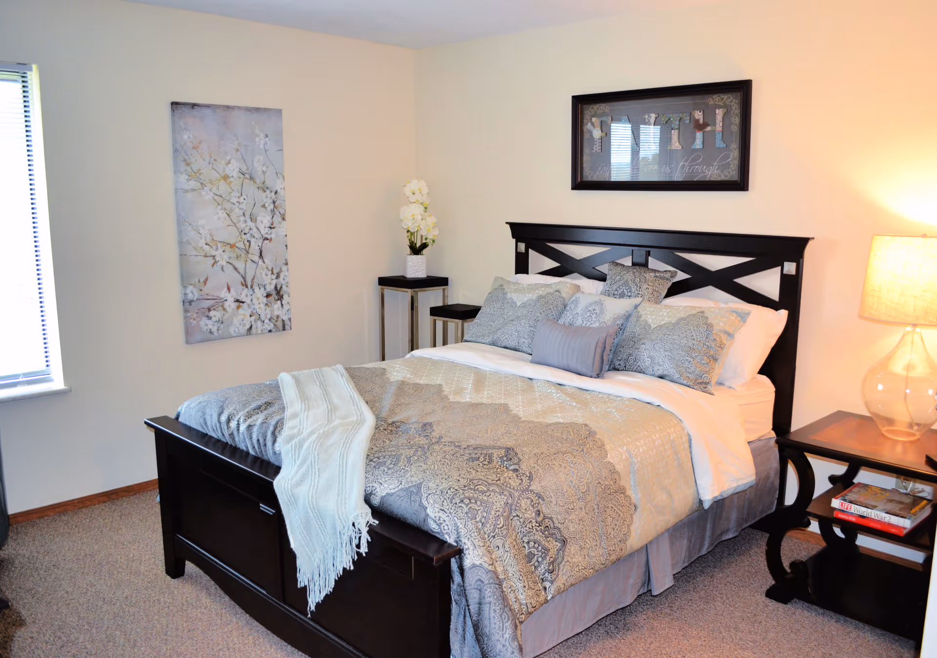 A neatly made bed with a dark wooden frame and patterned bedding in a bedroom. There are multiple pillows arranged on the bed, a light blue throw blanket draped over the footboard, and a bedside table with a lamp and books. A small table with a white orchid plant is placed in the corner, and two framed artworks hang on the walls. A window with blinds is visible on the left side.