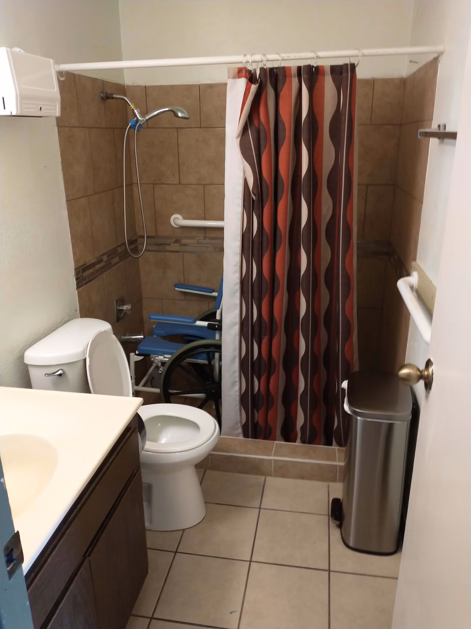 Small bathroom with beige tiled floor and walls, a white toilet with the lid open, a white sink countertop with dark wooden cabinet, a shower area with a red, brown, and beige patterned curtain partially drawn, a blue wheelchair inside the shower, a silver trash can, and grab bars on the walls for accessibility.