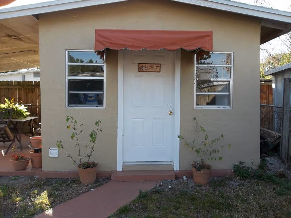 Exterior view of a small beige building with a white door labeled 'Laundry' under a red awning. There are two windows on either side of the door and potted plants placed near the entrance. A wooden fence and patio furniture are visible to the left.