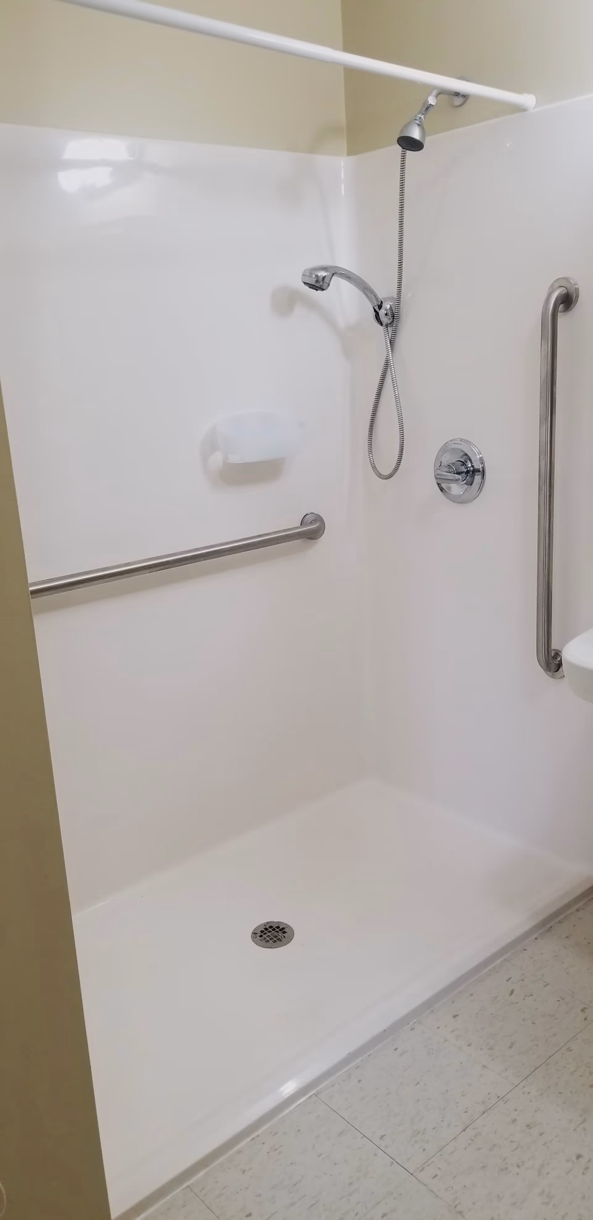 A clean, white shower stall with a handheld showerhead, two stainless steel grab bars, a soap dish, and a white shower curtain rod.