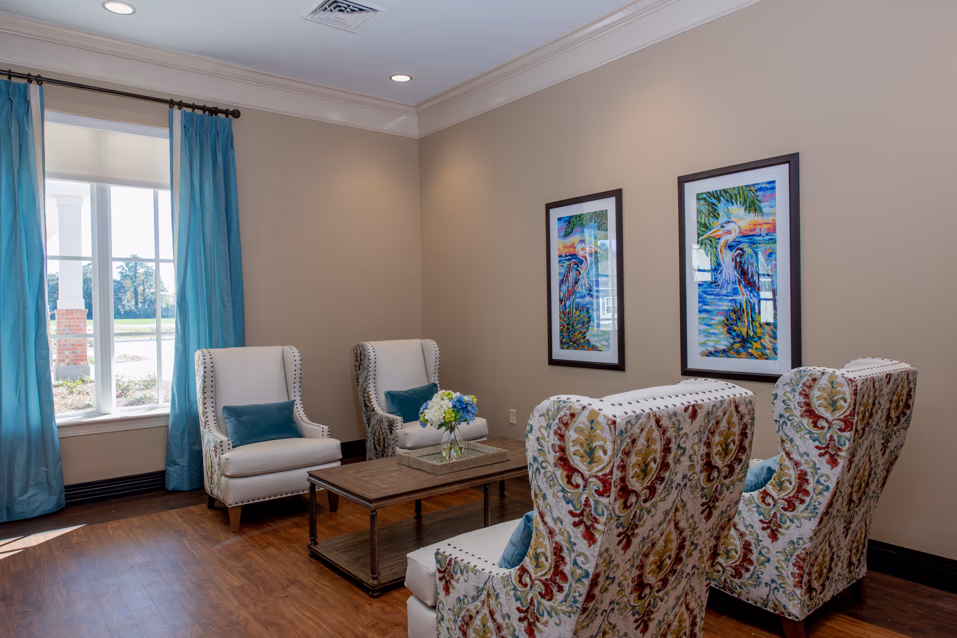 Bright sitting room with patterned wingback chairs, white armchairs, a wooden coffee table, blue curtains and framed artwork on the wall.