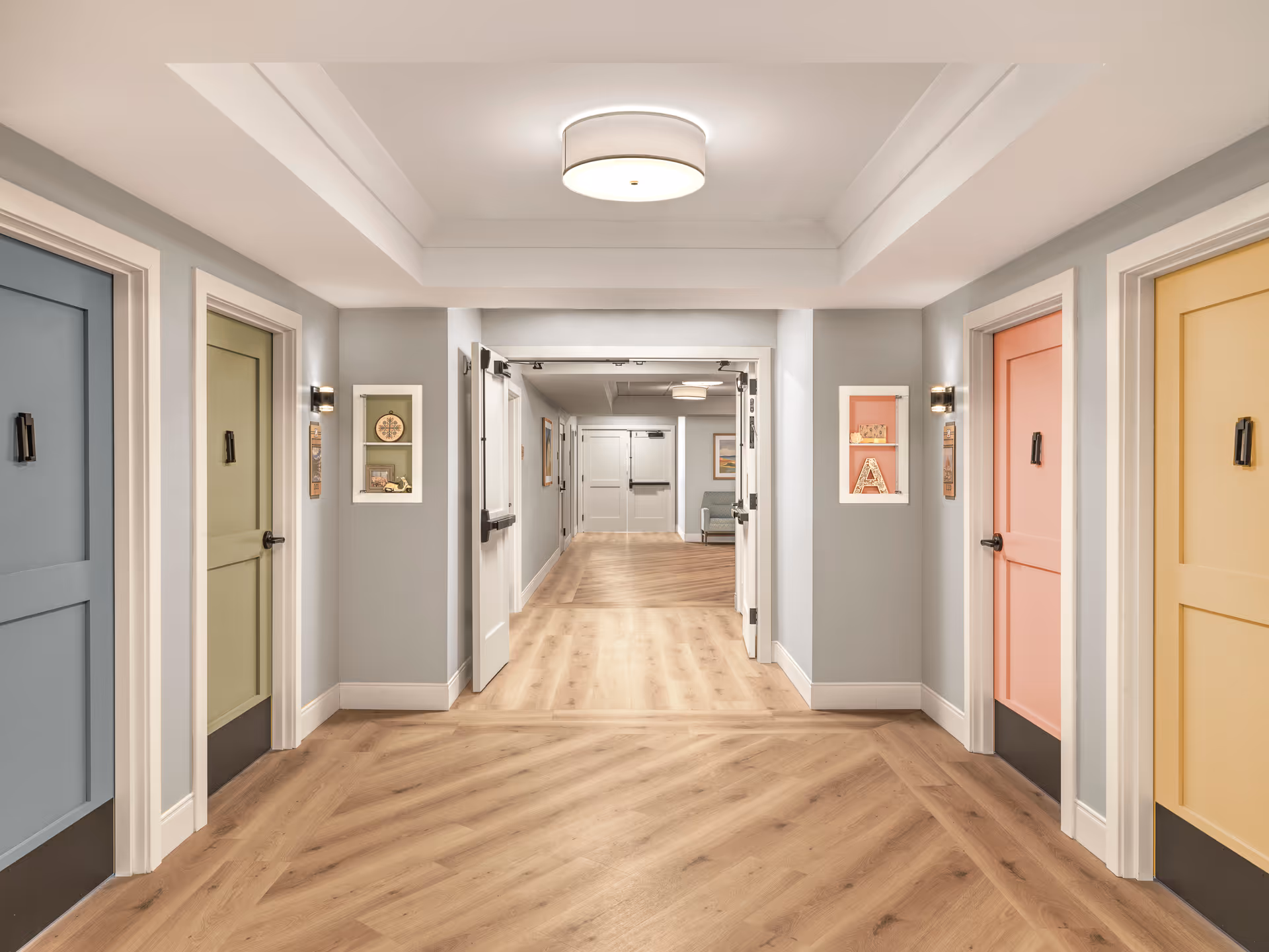 A bright, clean hallway in a senior living facility with light wood flooring and pastel-colored doors on both sides. The doors are painted in blue, green, pink, and yellow. The hallway has soft gray walls, white trim, and a modern ceiling light fixture. Small decorative shelves with items are mounted on the walls between the doors. Double doors at the end of the hallway lead to another area with seating visible.