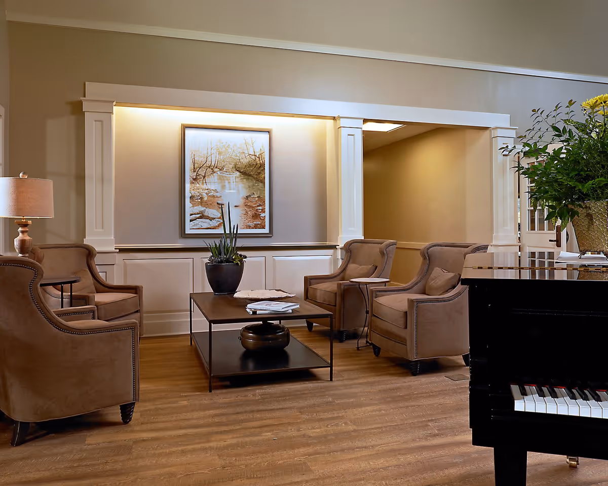 A cozy living room area with four brown upholstered armchairs arranged around a rectangular wooden coffee table with a plant and books on it. A framed painting of a river scene is mounted on the wall with soft lighting above it. To the right, part of a black piano with a flower arrangement on top is visible. The room has wooden flooring and beige walls.