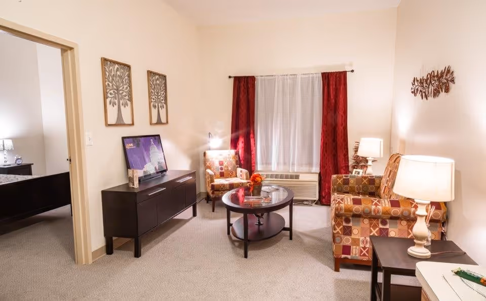A cozy living room with beige walls and carpet, featuring a patterned armchair and loveseat, a round coffee table with a small flower arrangement, a dark wooden TV stand with a flat-screen TV, two framed tree artworks on the wall, and a window with red curtains and white sheer curtains. A doorway on the left leads to a bedroom with a bed and nightstand visible.