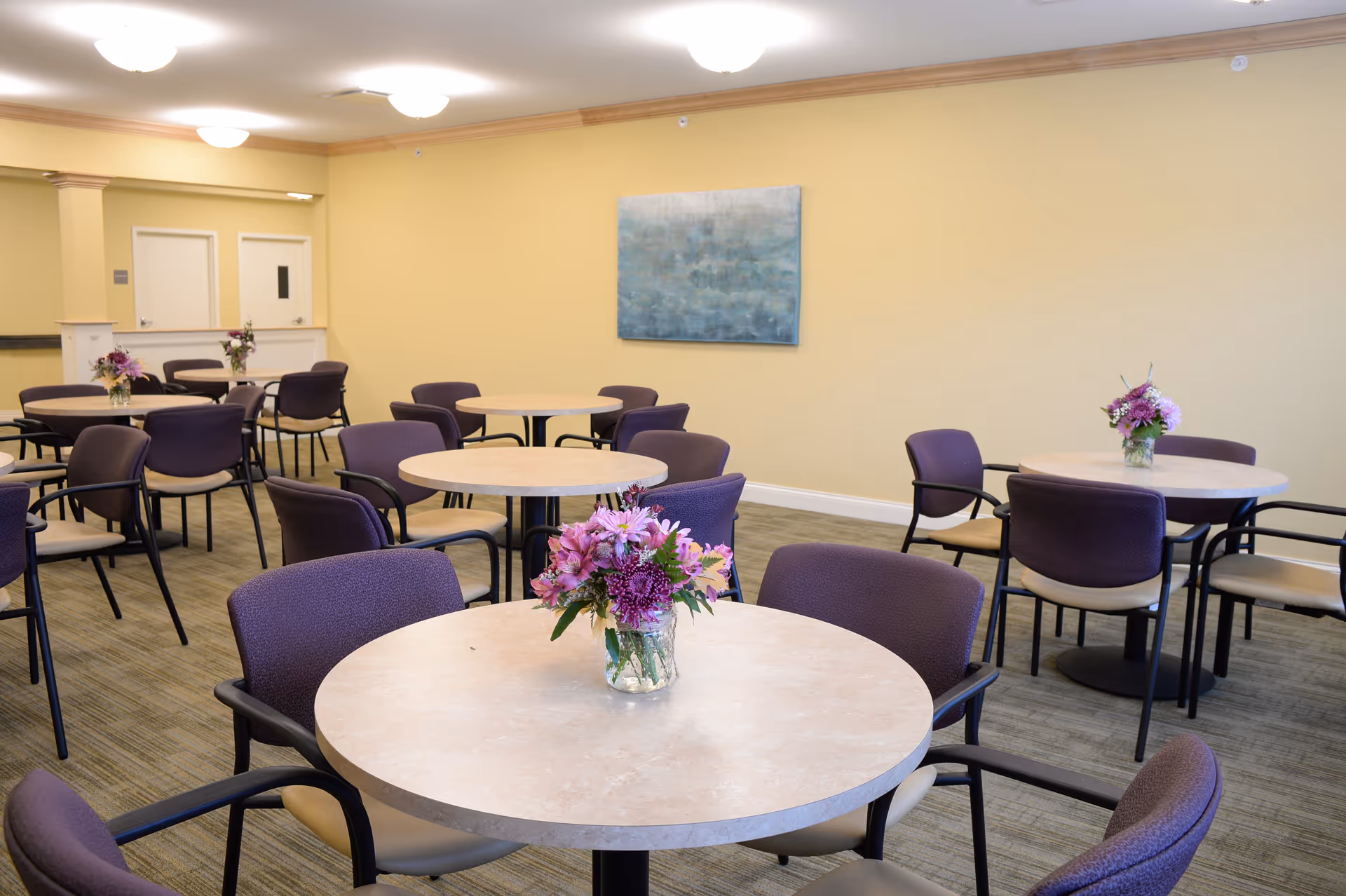 Well-lit dining room with round tables, purple chairs, and floral centerpieces.