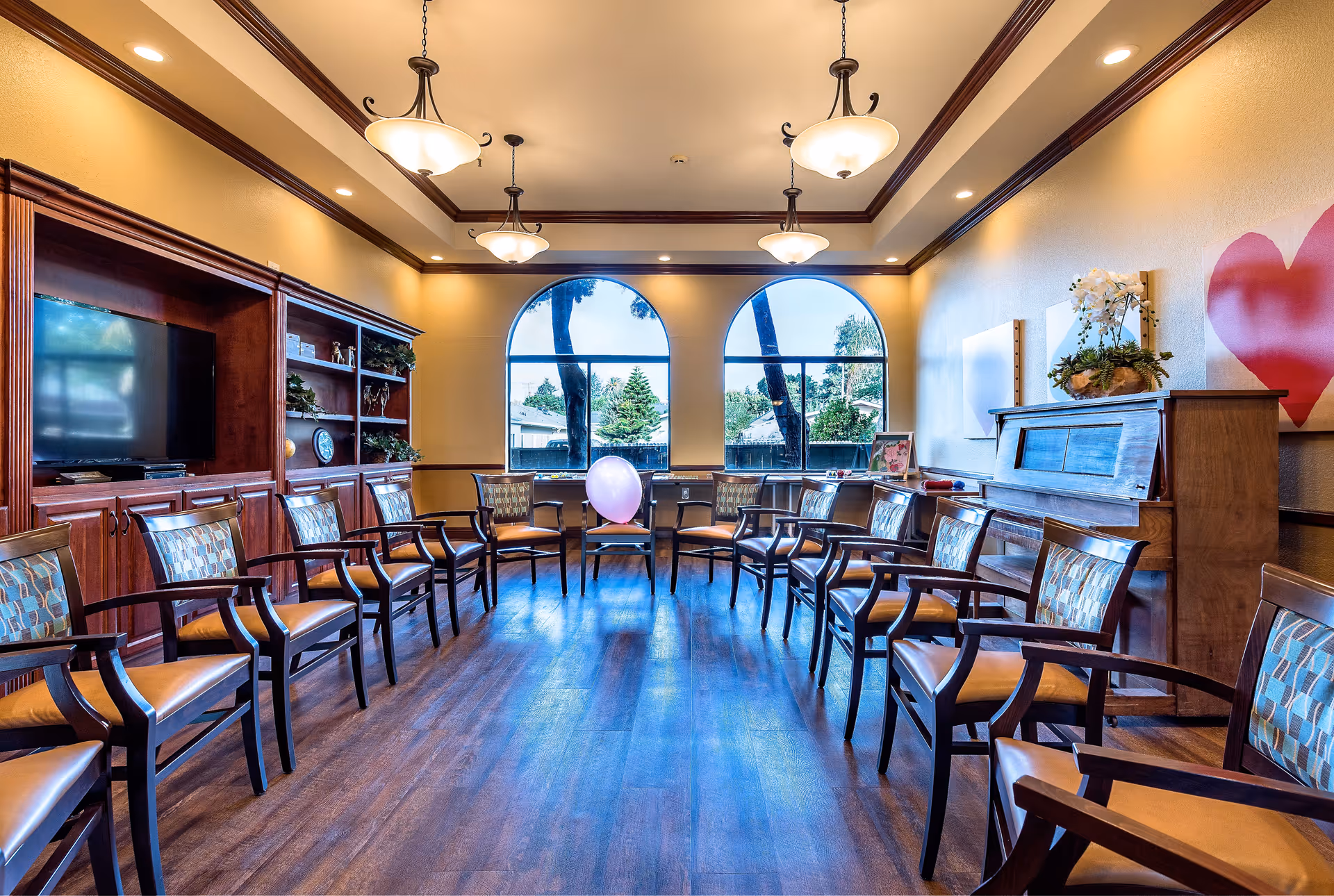 A spacious senior living common room with wooden chairs arranged in a large circle. The room features two large arched windows letting in natural light, a wooden piano with a flower arrangement on top, a built-in wooden entertainment center with a TV, and warm yellow walls with crown molding. A single purple balloon is placed on one of the chairs.