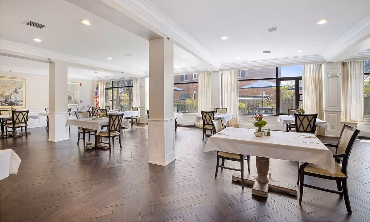 Bright, airy dining room with multiple tables draped in white linens and large windows overlooking an outdoor patio.
