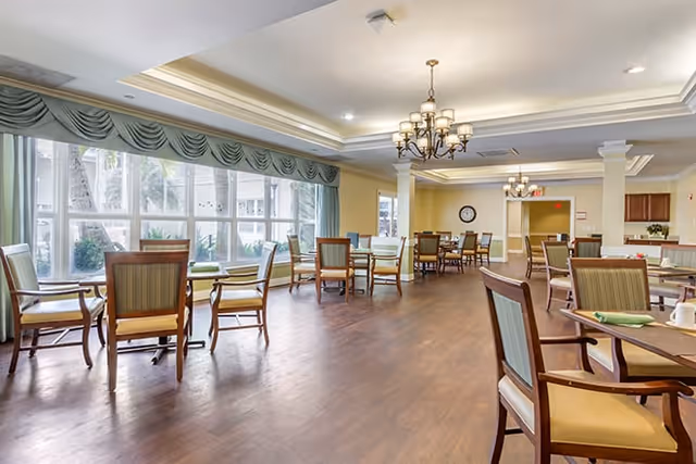 Bright senior living dining room with multiple tables and chairs, large windows with draped valances, chandeliers, and wood flooring.