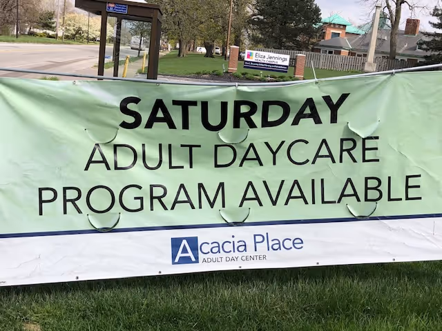 A large green and white banner displayed outdoors on grass, advertising a Saturday adult daycare program available at Acacia Place Adult Day Center. In the background, there is a street, trees, and a sign for Eliza Jennings Adult Day Care Center.